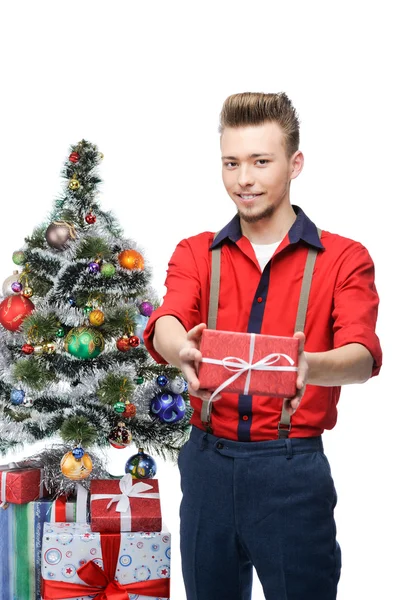 Joven hombre vintage sonriente sosteniendo regalo cerca del árbol de Navidad —  Fotos de Stock
