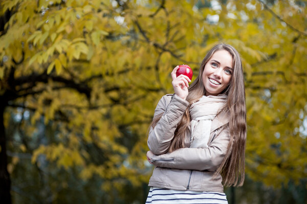 Young happy casual caucasian woman in autumn park smiling while holding red apple
