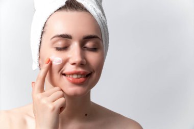 Kafasında havluyla güzel bir kadın gülümsüyor ve beyaz bir arka plana yüz kremi sürüyor.