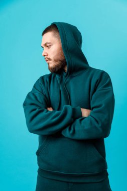 Yeşil eşofmanlı, sakallı yakışıklı genç adam kollarını mavi arka planda göğsüne katlamış. izole