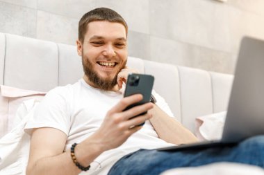 Evdeki kanepede dizüstü bilgisayarı ve telefonu olan genç adam.