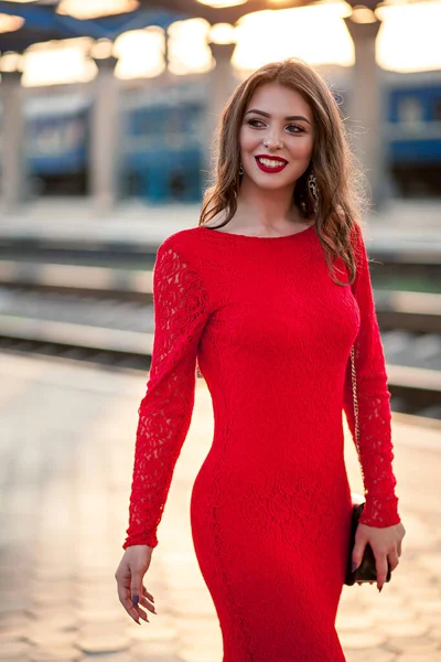 Uzun kırmızı elbiseli güzel bir kadın tren istasyonunun yanındaki platformda duruyor..