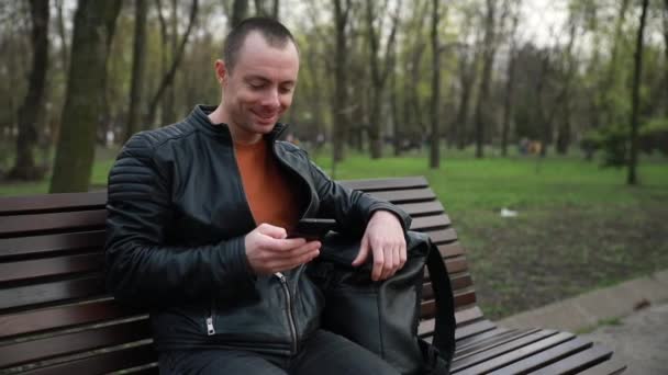 Business young man with a phone on a bench in a city park — Stok video