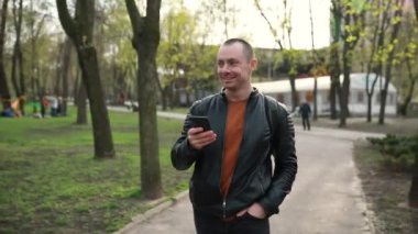 a handsome young cheerful man walks through the park with a phone in his hands. Spring