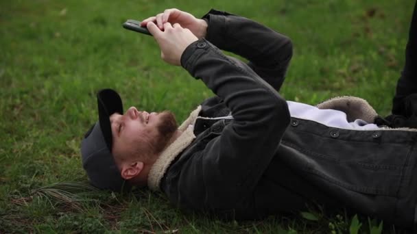 Young man lies on green grass with phone — Wideo stockowe