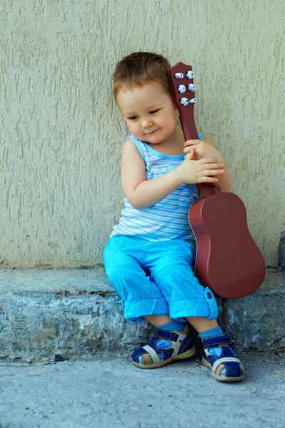 Милый ребенок мальчик с гитарой, сидя против бетонная стена