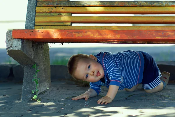 Roliga busiga baby boy kikar ut från under bänken. utomhus — Stockfoto