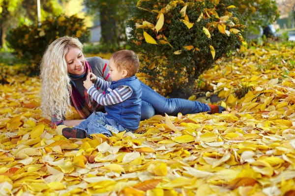 Attractive curly mother and son sitting on the carpet of fallen leaves in a — Stock Photo, Image