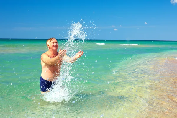 Šťastný zralý muž šplouchání vody na pláži — Stock fotografie