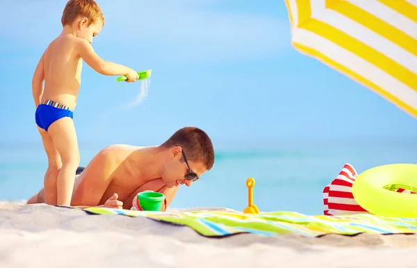 Playful son strews sand on father, colorful beach — Stock Photo, Image