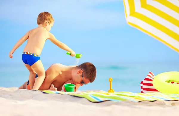 Playful son strews sand on father, colorful beach — Stock Photo, Image