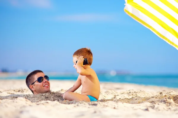 Ragazzo felice che parla al telefono, testa di padre nella sabbia — Foto Stock