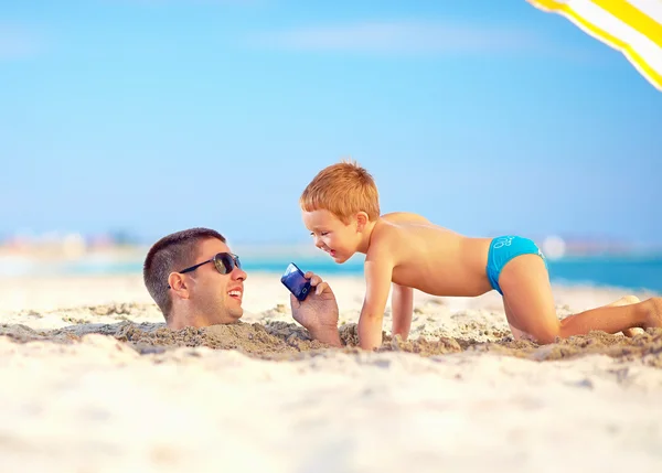 Boldog apa és fia együtt, beszél szúró telefon a strandon — Stock Fotó