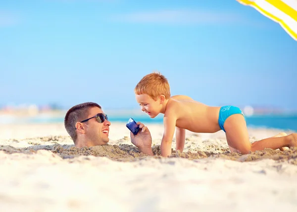 Gelukkig vader en zoon praten over slimme telefoon samen, op het strand — Stockfoto