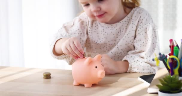 幼い頃から子供を救うこと 幸せな幼児の赤ん坊の女の子は貯金箱に硬貨を落とす あなたの子供にお金箱で予算を計画するように教える — ストック動画