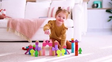 Sevimli küçük kız renkli tahta bloklarla oynuyor, güneşli bir günde evde halının üzerine oyuncak bir kale inşa ediyor.