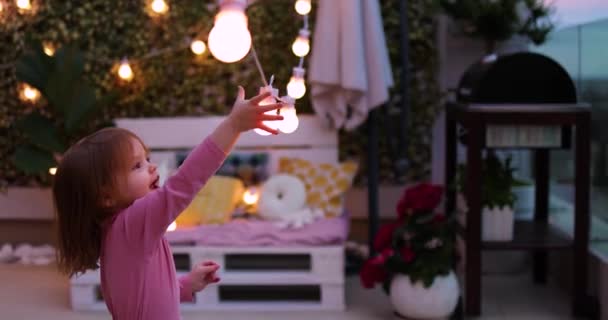 明るい幼児の女の子は夏の夜に屋上のパティオで楽しみ パーティー電球の文字列で遊んで — ストック動画