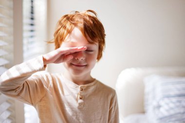 cute redhead young boy rubbing his eyes in the morning, waking up in sunlit bedroom