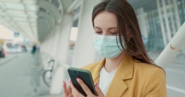 Tıbbi maskeli genç iş kadını şehir merkezinde oturuyor ve modern akıllı telefonlarda internete takılıyor. Açık havada moda sarı ceketli bir kadın.. 