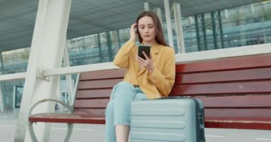 Elinde bavuluyla bankta oturan ve modern akıllı telefon kullanan güzel bir kadın. Sarı ceketli bayan dışarıda uçağı bekliyor..