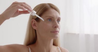 Senior woman using anti wrinkle serum in the bedroom. Naked mature woman standing holding pipette of lactic acid applying on face smiling relaxed. Beauty concept 