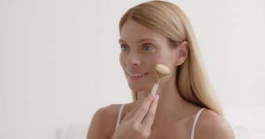 Portrait of senior mid aged woman of 50s years old massaging face skin using natural stone trendy jade quartz rolling device for detox of perfect skin. Spa traditional technique skincare ads 