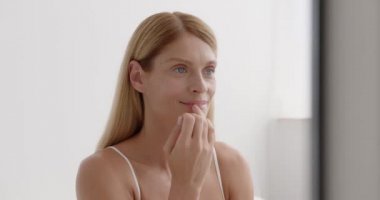 Close-up portrait of lovely blonde middle aged lady smooth skin using lip balm isolated over white bedroom background 