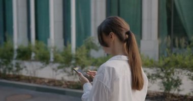 Smiling young woman wearing in white shirt walking around modern street and using smartphone. Communication, social networks, online shopping concept 