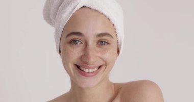 Portrait of caucasian woman without makeup wrapped in towel smiling sincerely to camera after using facial skin care cream. Lady makes skin care procedures. Spa, daily treatment and wellness concept 