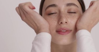 Portrait of beautiful young woman applying face cream and smiling sincerely in studio with white background. Vertical video of happy caucasian female model