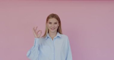 Portrait of satisfied beautiful young woman with blonde hair in casual blouse standing, looking at camera showing Ok sign gesture, indoor studio shot isolated on pink background 