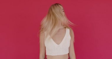 Young woman with long hair wearing white t shirt waving hair, looking seriously, expressing calm emotion isolated on pink background indoors 