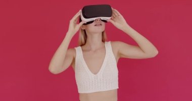 Amazed woman using VR app headset helmet to play simulation game. Watching virtual reality 3d video. Isolated on pink background in studio. Girl in VR goggles looking around 