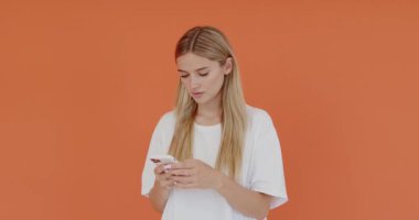 Positive optimistic female using smartphone with happy facial expression, hearing pleasant news, wearing white casual style t shirt. Indoor studio shot isolated on orange background
