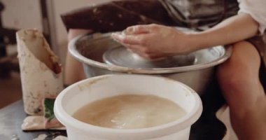 Woman potter pouring water while working on potters wheel. Girl making ceramic pot from clay in pottery workshop. Traditional art concept, creative workshop and masterclass