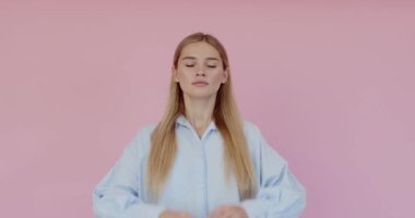 Portrait of peaceful cute woman wearing casual style shirt holding fingers in mudra gesture and meditating with closed eyes, yoga practice. Indoor studio shot isolated on pink background 