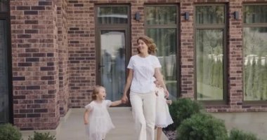 Tatlı kızlarının el ele tutuşan ve tuğla evin yanında yürüyen hoş kadının uzun bir görüntüsü. Mutlu aile kavramı