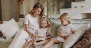 Genç anne ve kızlarından oluşan mutlu bir aile kitap okuyor, rahat bir şekilde kucaklaşıyor ve rahat bir oturma odasında kanepede oturuyor.