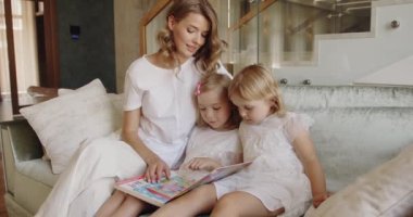 Sevimli anaokulu kızları ellerinde kitap okuyarak anneleriyle birlikte kanepede oturuyorlar. Aile zamanı kavramı 