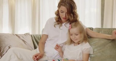 Küçük bir çocukla rahat koltukta oturan gülümseyen genç anne. Mutlu anne evde küçük bir kızla kitap okurken eğleniyor. Annelik, çocuk bakımı kavramı 