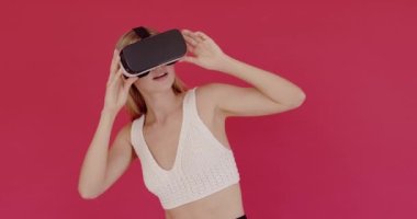Sarışın kız, simülasyon gerçekçi oyunu oynamak için kulaklık uygulaması kullanıyor. Sanal gerçeklik videosu izliyorum. VR gözlüklü genç kız pembe stüdyo arka planında izole edilmiş. 