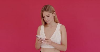 Akıllı telefon kullanan kadın portresi, sosyal ağları kontrol eden, ifade odaklı olan, beyaz günlük kıyafet giyen. Pembe arka planda kapalı stüdyo çekimleri 