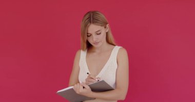 Kağıt deftere yazı yazan akıllı kadın yapılacaklar listesini tamamlıyor ya da çalışıyor. Pembe arka planda kapalı stüdyo çekimleri 