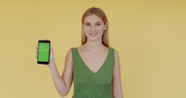 Güzel görünümlü, krom tuşlu akıllı telefon gösteren, yeşil ekrana işaret eden, güvenli bir şekilde sarı arka planda izole edilmiş en iyi fiyat sunan genç bir kadın. 