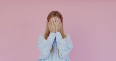 Genç, komik, güzel bir kadın elleriyle yüzünü gizliyor ve gülümsüyor. Mutluyum, gülüyorum. Pembe arka planda izole edilmiş olumlu duygularla kız kamuflajı ve yüzünü göster 