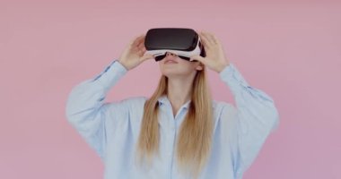 VR kulaklık takan, elleriyle havaya dokunan, heyecanlı ifadeyle sanal oyun oynayan kadın portresi. Pembe arka planda kapalı stüdyo çekimleri 