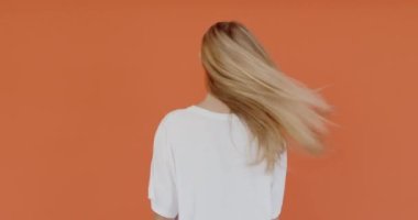 Uzun saçlı, beyaz tişörtlü genç bir kadın kameraya doğru arkasını dönüyor, ciddi görünüyor, turuncu arka planda izole edilmiş sakin duygularını ifade ediyor. 