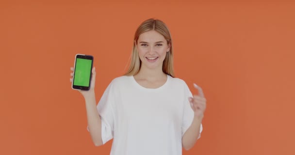 緑の画面を指して クロマキーモックアップ付きスマートフォンを示す良い外観を持つクールな若い大人の女性は 自信を持ってオレンジの背景に隔離された最高の取引価格を提供屋内 — ストック動画