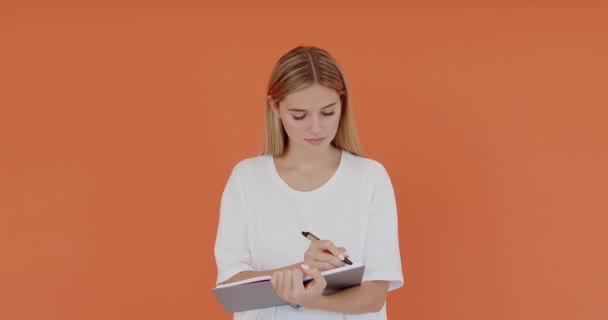 紙のノートにメモを書き 創造的なアイデアを書き 将来の計画とやるべきリストを作成する幸せな美しい学生の女の子は インスピレーションを探しています オレンジの背景に隔離された屋内スタジオショット — ストック動画