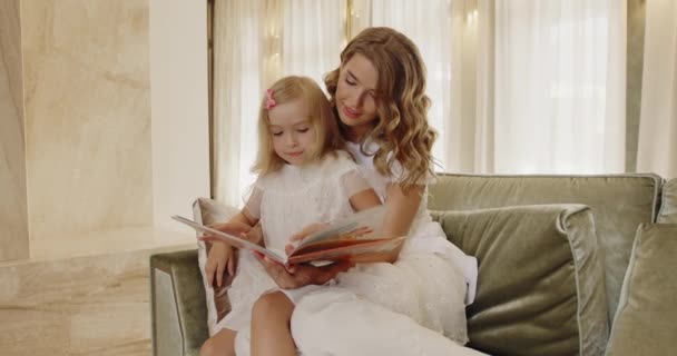 かわいい金髪の就学前の娘が一緒にソファに座ってお母さんとおとぎ話を読んで本を保持しています 家族の時間概念 — ストック動画